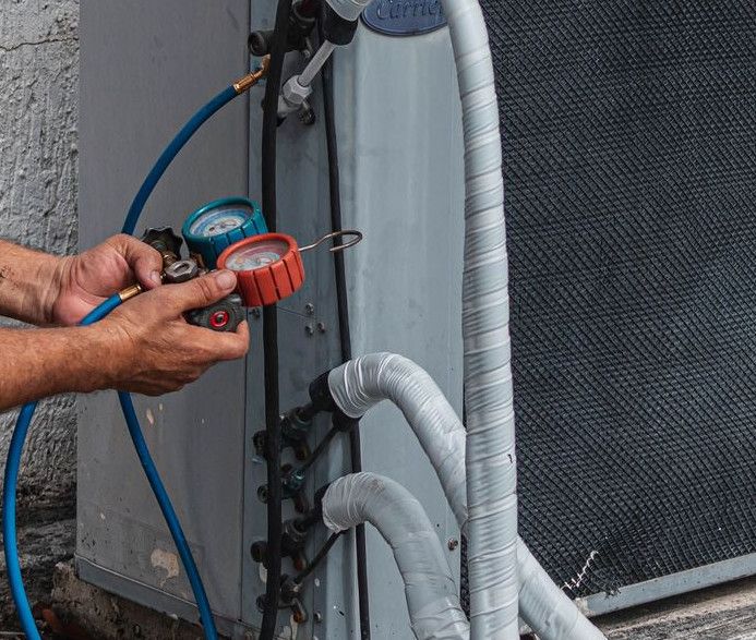 Technician repairing an air conditioner unit outdoors, wearing a facemask and using a manifold gauge.
